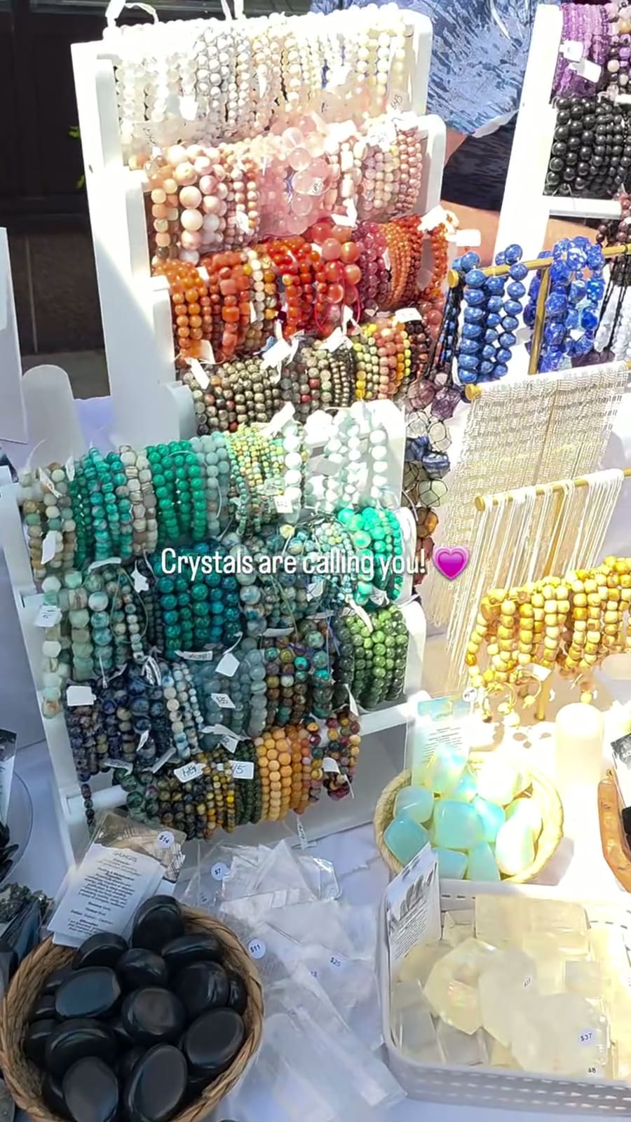 Racks of beaded bracelets arranged by colour, running from rose and carnelian through green and turquoise to lapis and gold. Printed across the photograph: “Crystals are calling you!”.