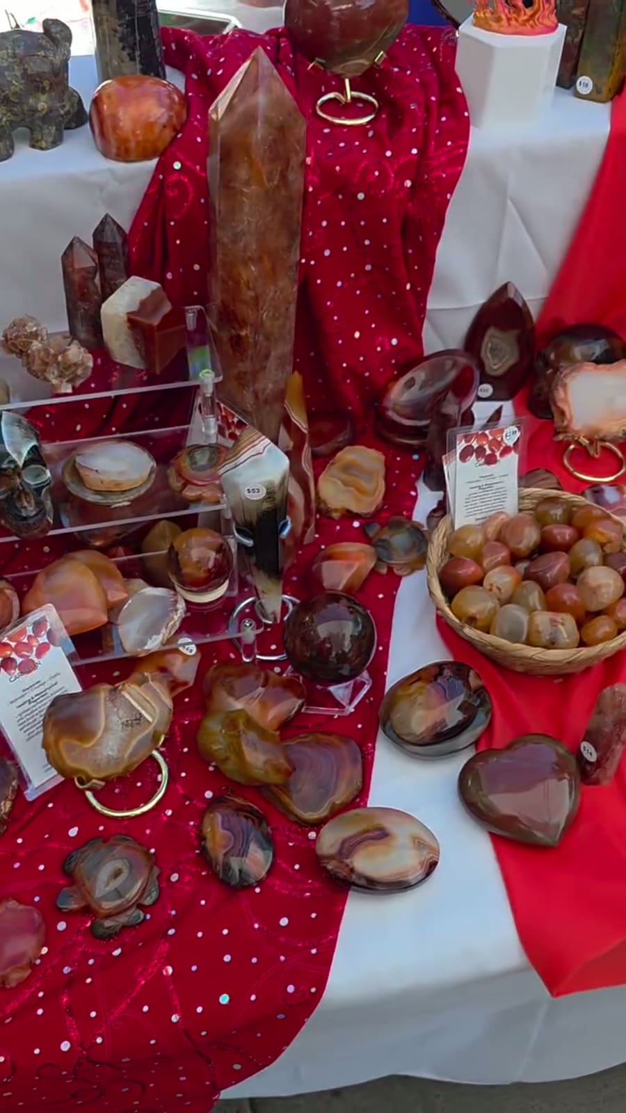 A table under red and white cloth covered in carnelian and agate — a tall polished point, spheres on stands, hearts, slices and a basket of tumbles.