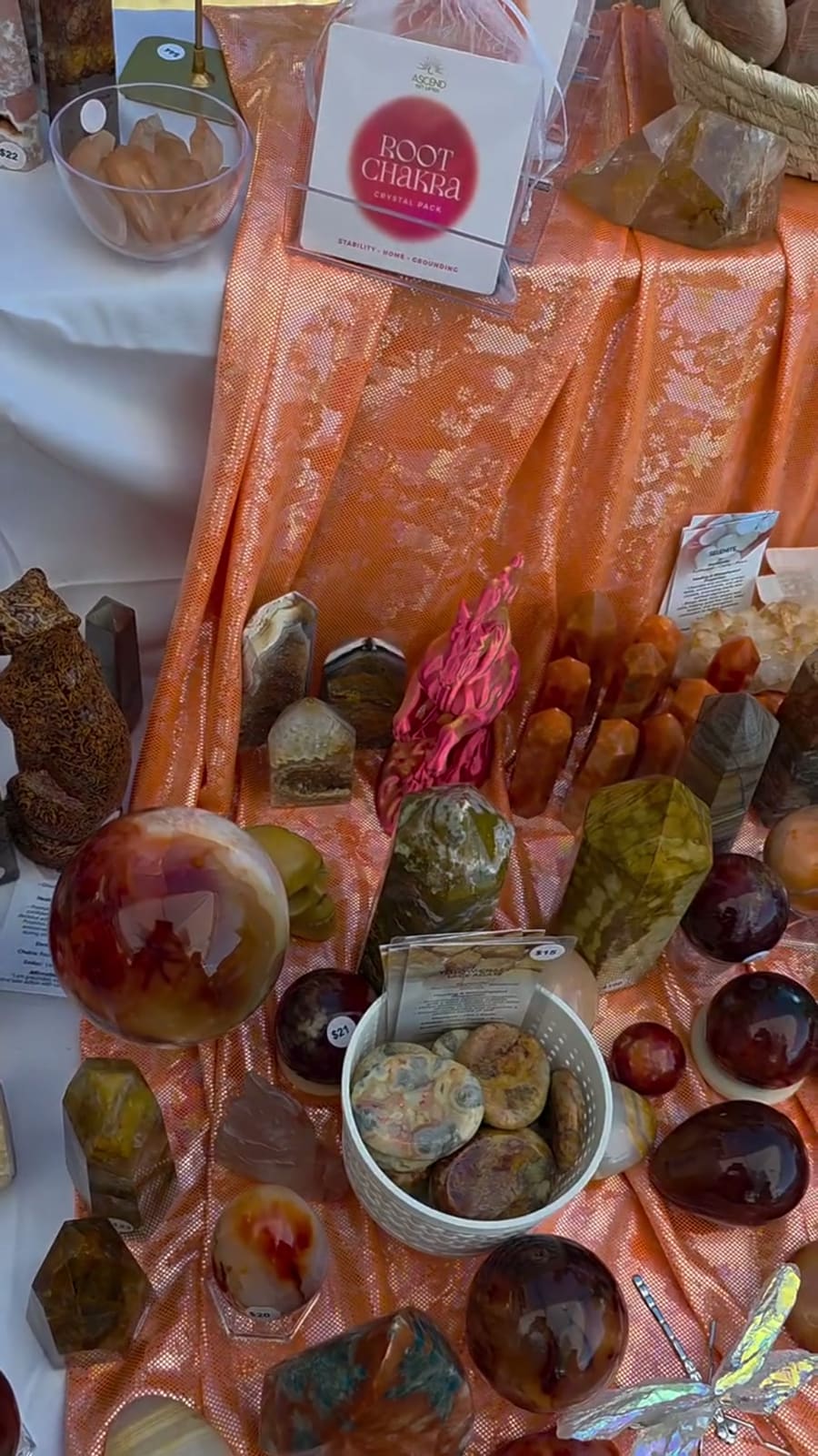 Coral-orange cloth crowded with carnelian points, a large banded sphere, a colander of jasper tumbles and a hanging Root Chakra crystal pack.