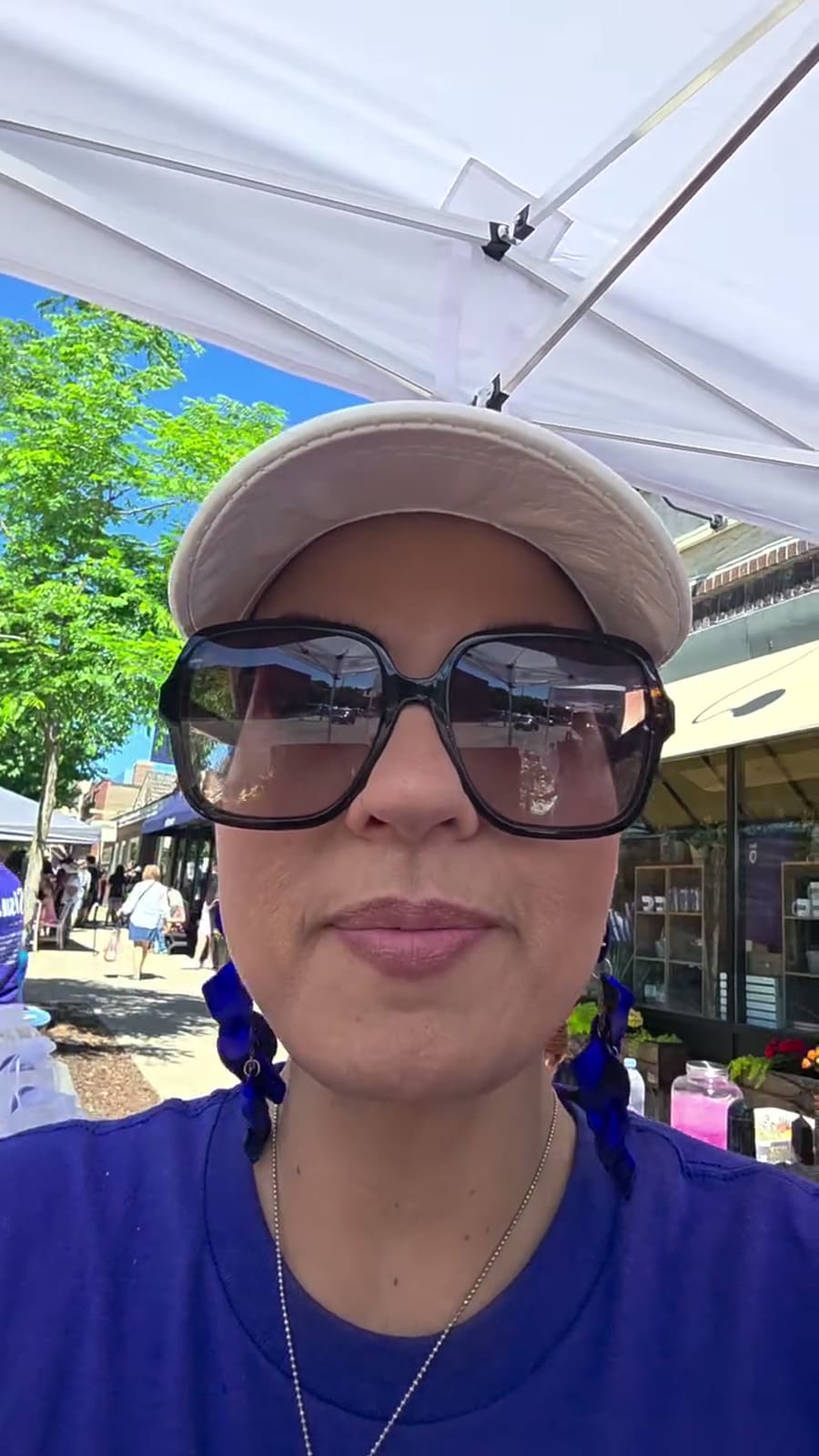 Sandra under the white canopy of the stall, in sunglasses and a cap, with the fair street and the shopfronts behind her.