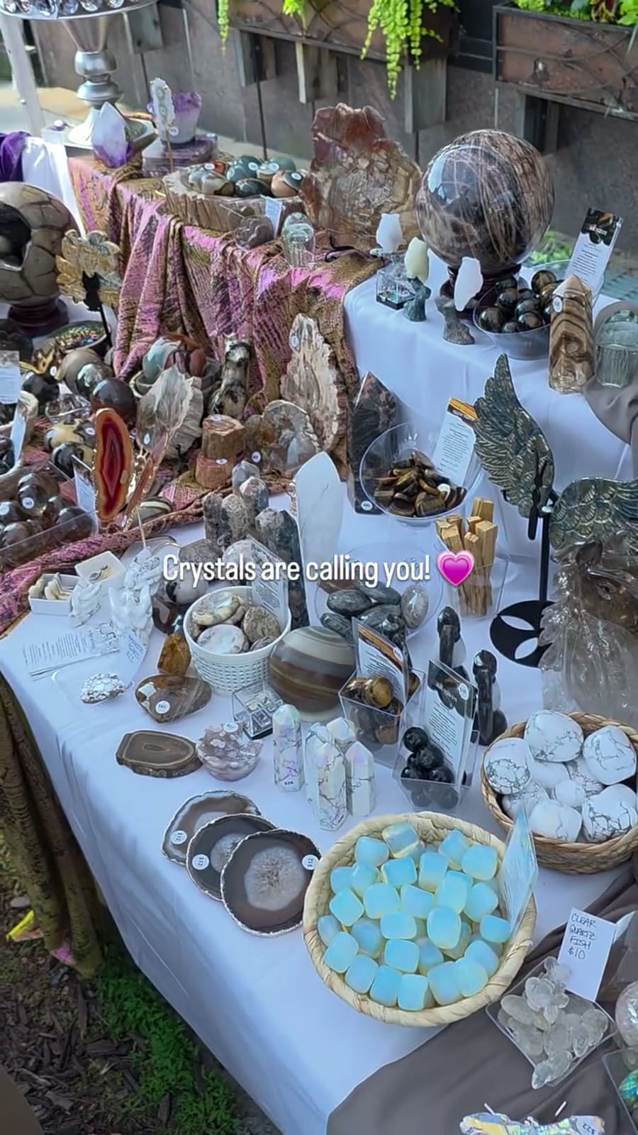 A white-clothed table of petrified wood, a large brown sphere on a stand, agate slices, palo santo and a basket of pale opalite cubes. Printed across the photograph: “Crystals are calling you!”.