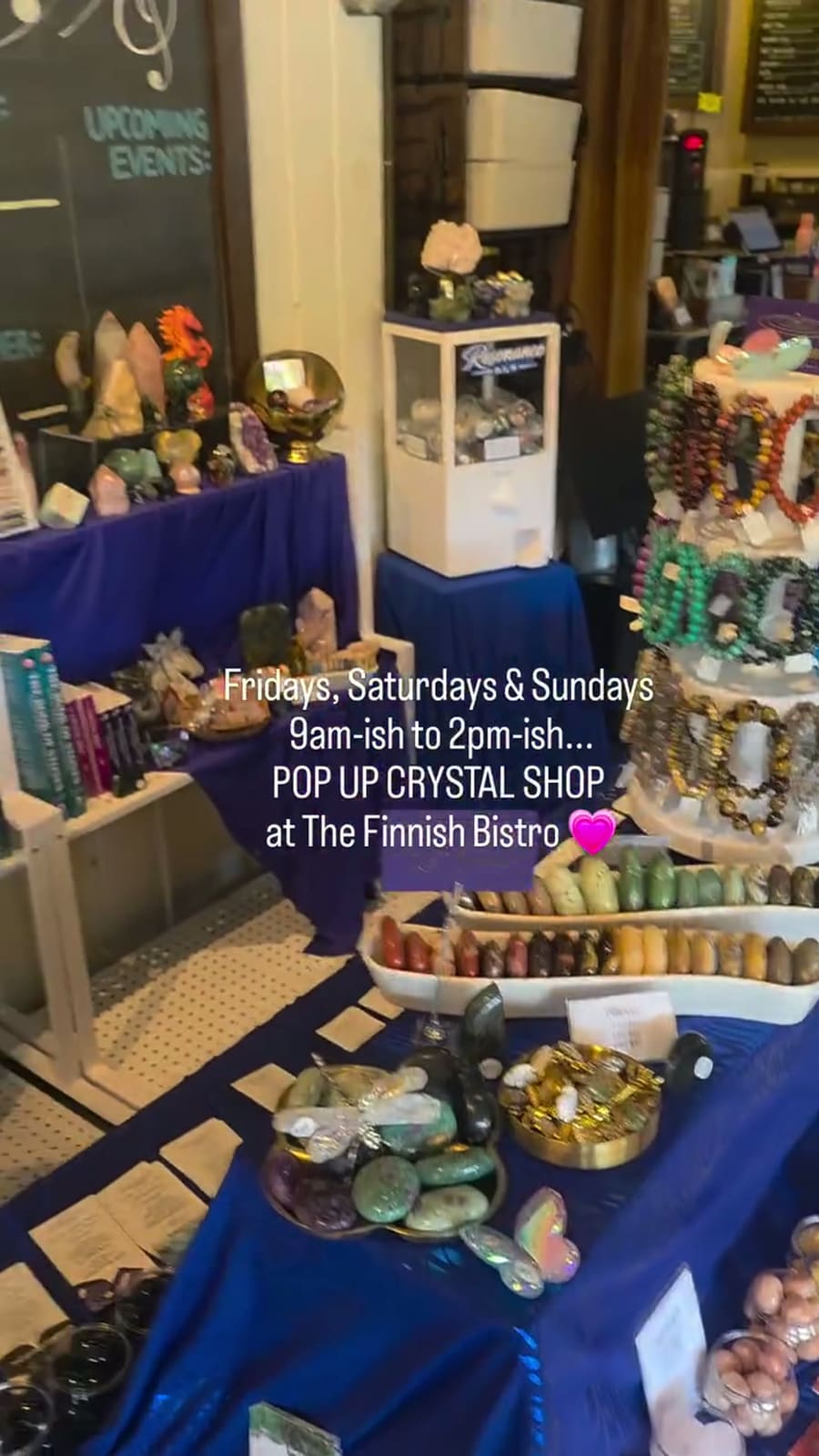The stall set up inside The Finnish Bistro: indigo-draped tables of towers and spheres, a tiered stand of beaded bracelets, and the cafe’s chalkboard behind. Printed across the photograph: “Fridays, Saturdays & Sundays, 9am-ish to 2pm-ish… POP UP CRYSTAL SHOP at The Finnish Bistro”.